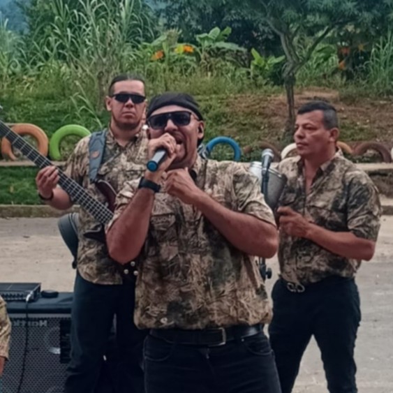 Grupo musical La Banda de la locura interpretando en vivo al aire libre. Los integrantes visten camisas camufladas en tonos marrón y verde, mientras el vocalista canta con pasión al centro, rodeado de músicos con bajo, caja y acordeón. Al fondo, vegetación tropical y llantas de colores decoran el entorno.Grupo musical La Banda de la locura interpretando en vivo al aire libre. Los integrantes visten camisas camufladas en tonos marrón y verde, mientras el vocalista canta con pasión al centro, rodeado de músicos con bajo, caja y acordeón. Al fondo, vegetación tropical y llantas de colores decoran el entorno.