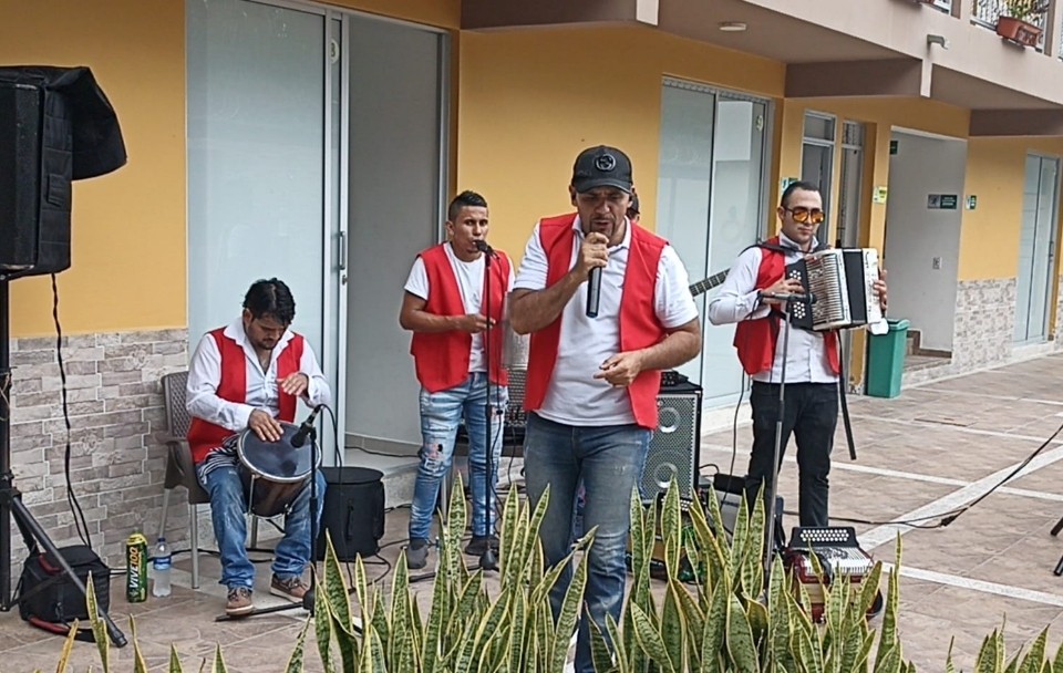 El grupo musical 'La Banda de la locura' realiza una presentación en un espacio abierto frente a un edificio. Cuatro integrantes, vestidos con chalecos rojos y camisas blancas, interpretan música vallenata con micrófono, caja, guacharaca y acordeón. Plantas decorativas en primer plano